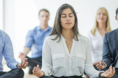 Una mujer que practica bienestar y salud mental meditando frente a un grupo de empresarios.