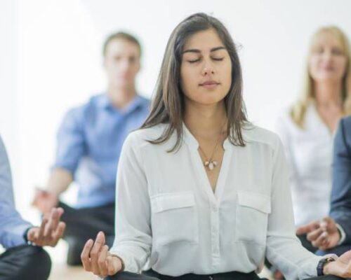 Una mujer que practica bienestar y salud mental meditando frente a un grupo de empresarios.