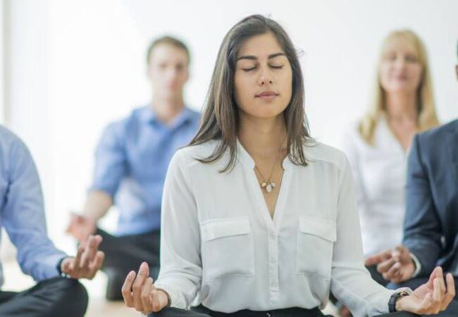 Una mujer que practica bienestar y salud mental meditando frente a un grupo de empresarios.